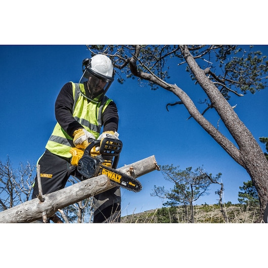 A person wearing safety gear is using a DEWALT chainsaw to cut a tree branch outdoors. The sky is clear and trees are visible in the background.