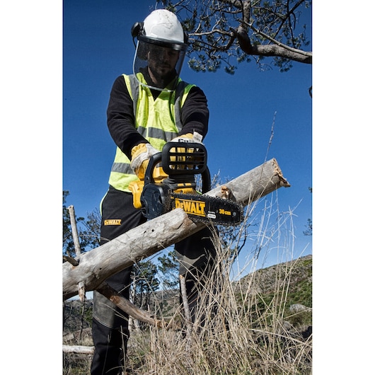 A person wearing safety gear and a high-visibility vest is using a DEWALT DCM565P1 chainsaw to cut a branch outdoors in a natural setting.
