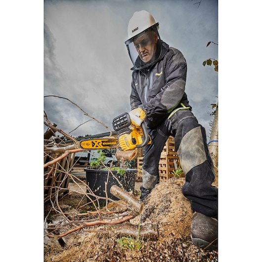 A person wearing protective gear is outdoors and using a DEWALT DCM565P1 battery-powered chainsaw to cut tree branches.