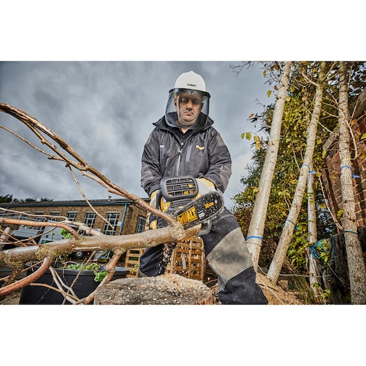 A person wearing protective gear and a visor is using a DEWALT DCM565P1 cordless chainsaw to cut a tree branch outdoors. The scene shows safety equipment and a building in the background.