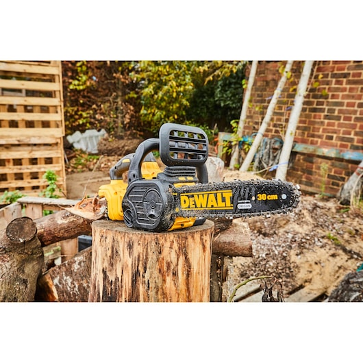 A DEWALT cordless chainsaw with a 30 cm blade is placed on a tree stump outdoors, surrounded by logs, with a brick wall and wooden pallet in the background.