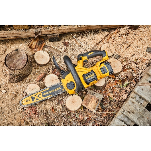 A yellow and black DEWALT cordless chainsaw lies on the ground among chopped wood logs and wood shavings outdoors.