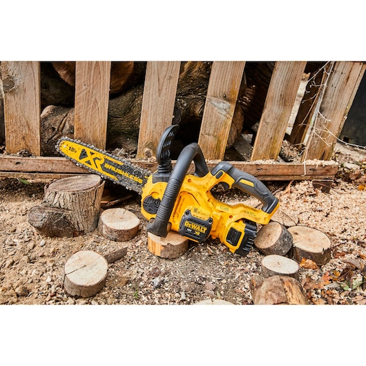 A yellow DEWALT XR Brushless 18V cordless chainsaw is placed on the ground among cut wooden logs and sawdust, with a wooden fence and stacked logs in the background.
