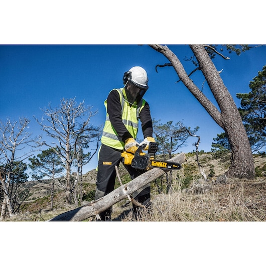 A person wearing safety gear, including a helmet, gloves, ear protection, and a yellow reflective vest, is using a DEWALT DCM565P1 cordless chainsaw to cut a tree branch outdoors in a forested area under a blue sky.