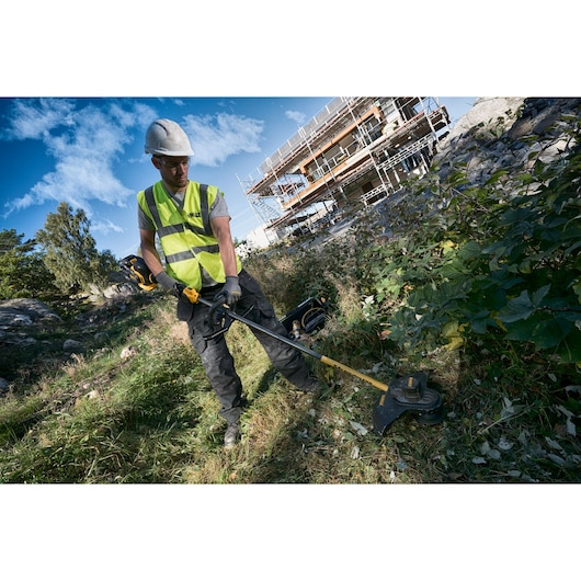 Worker using a DEWALT brush cutter outdoors, wearing safety gear, with a building site visible in the background.
