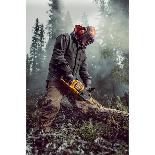 A person wearing protective clothing and a helmet is using a DEWALT DCM575X1 chainsaw to cut through a fallen tree in a forest. Wood chips are flying as the chainsaw operates.
