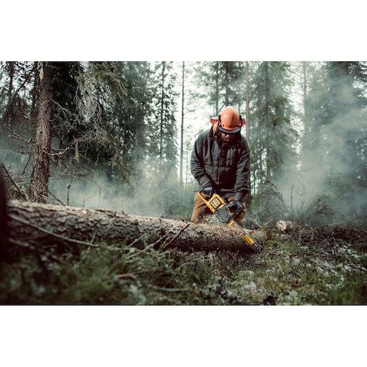 A person wearing protective gear operates a DEWALT chainsaw (DCM575X1), cutting a fallen tree trunk in a forest.