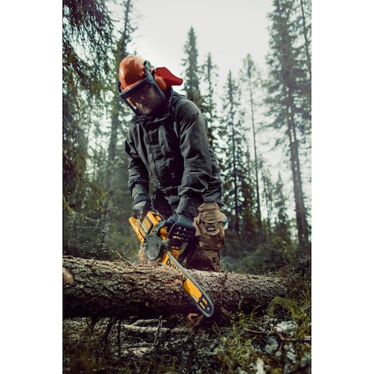 A person wearing safety gear uses a DEWALT DCM575X1 cordless chainsaw to cut a tree trunk in a forest.