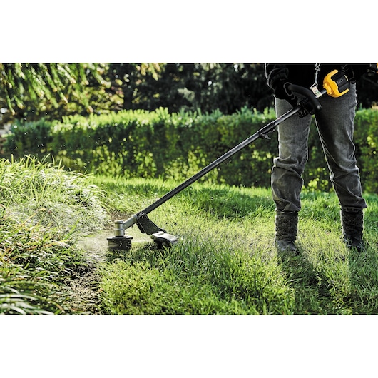 Person using a DEWALT cordless grass trimmer to cut tall grass in a garden area.