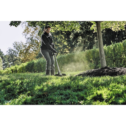A person is using a DEWALT cordless grass trimmer to cut grass around a tree in a garden or park. The photo is taken outdoors in daylight, showing green grass, trees, and hedges.