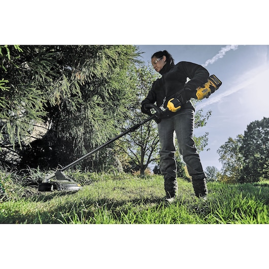 A person using a DEWALT cordless grass trimmer (model DCMAS5713X1) to trim tall grass in an outdoor garden area. The person is wearing dark clothing and gloves and their face is blurred. Trees and greenery are visible in the background.