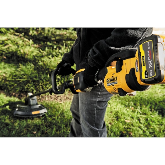 A person using a DEWALT 54V XR FLEXVOLT cordless brushless string trimmer to cut grass in a garden. The close-up shows the DEWALT branding and the battery attached to the tool.