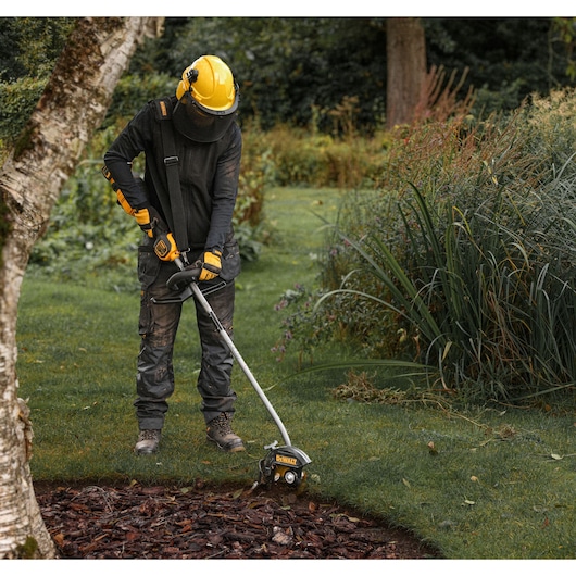 DEWALT DCMASED9N, DCMASED9N-XE edger in use for garden work. Person wears protective gear outdoors.