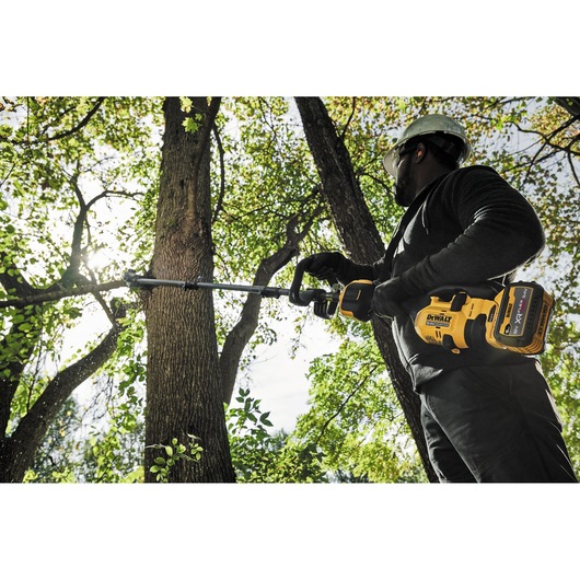 A person wearing safety gear uses a DEWALT cordless pole saw to prune tree branches outdoors.
