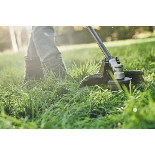 Close-up view of a string trimmer being used to cut grass outdoors. The person operating the trimmer is partly visible, focusing on their legs and boots in a grassy area.