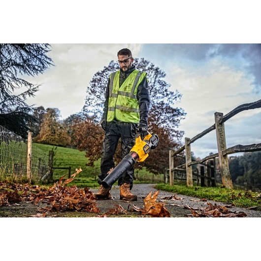A person wearing a high-visibility safety vest is using a yellow and black DEWALT cordless leaf blower (model DCMBA572X1) to clear fallen leaves from a path in an outdoor setting. Wooden fencing and autumn trees are visible in the background.