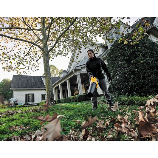 A person uses a DEWALT leaf blower to clear fallen leaves from a grassy yard in front of a large house.