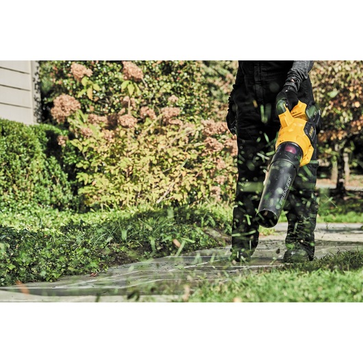A person uses a DEWALT cordless leaf blower to clear grass clippings from a garden path, with greenery and plants in the background.