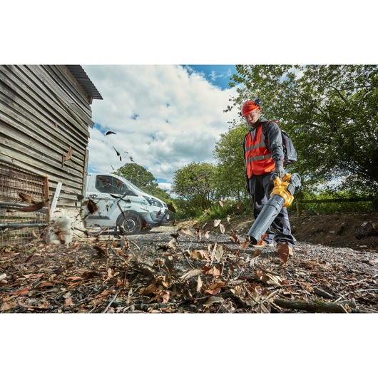 A person wearing safety gear and a red vest uses a DEWALT leaf blower outdoors to clear leaves and debris from a gravel area. There is a white van, a wooden building, and trees in the background.