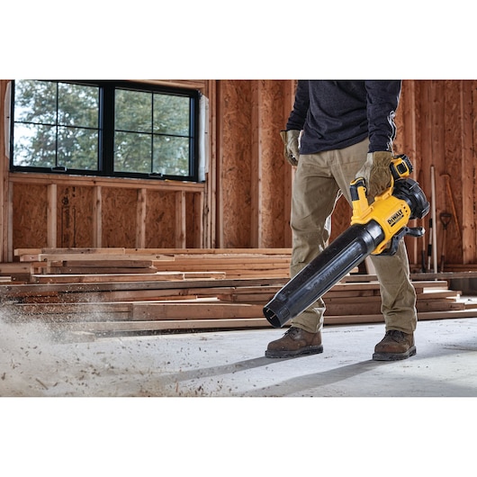 A person using a DEWALT cordless blower (model DCMBL562N) to clear dust and debris from the floor inside a wooden