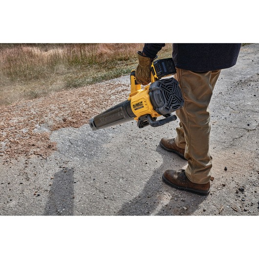 A person wearing gloves and work boots uses a DEWALT cordless leaf blower to clear debris from a paved outdoor surface.