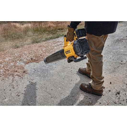 A person using a DEWALT battery-powered leaf blower to clear debris from a paved surface outdoors.
