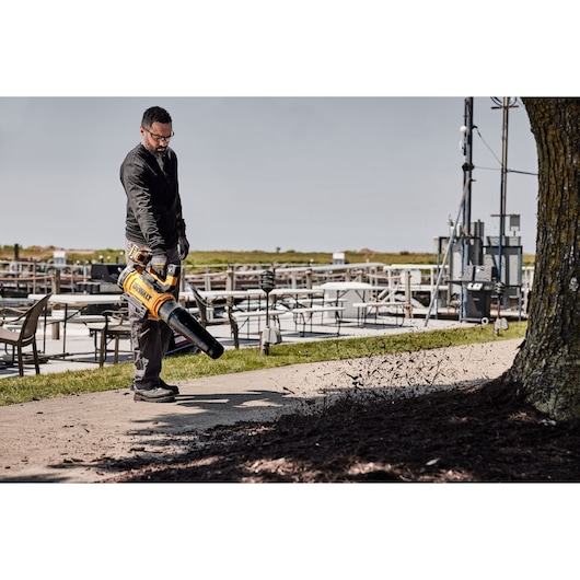 A person using a DEWALT cordless leaf blower (DCMBL777N) to clear debris from a sidewalk in an outdoor area with tables and