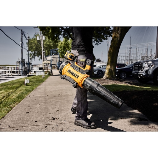 A person holding a DEWALT cordless leaf blower, model DCMBL777N, using it to clean debris from a sidewalk in an outdoor