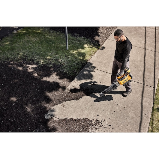 A person using a DEWALT cordless leaf blower to clear debris from a sidewalk next to grass and mulch.