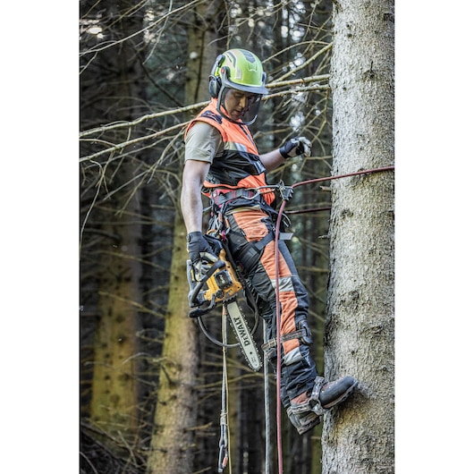 A person equipped with safety gear and helmet is using a DEWALT chainsaw (model DCMCST635X1) while climbing a tree in a