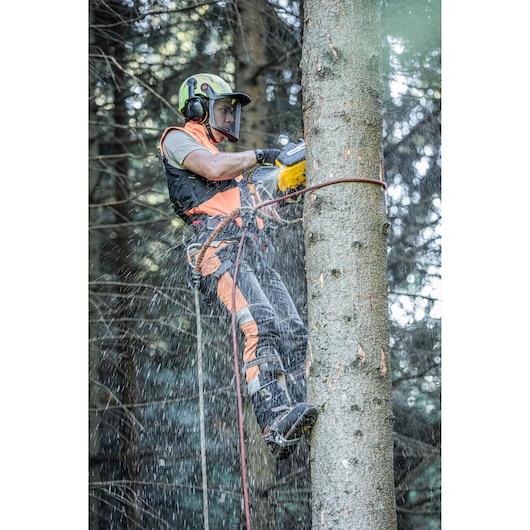 A person wearing safety gear, including a helmet and ear protection, uses a yellow DEWALT chainsaw to cut a tree trunk while