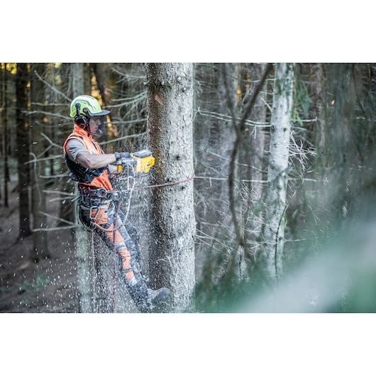 A worker wearing safety equipment uses a DEWALT cordless chainsaw to cut a tree in a forest. The worker is secured with a