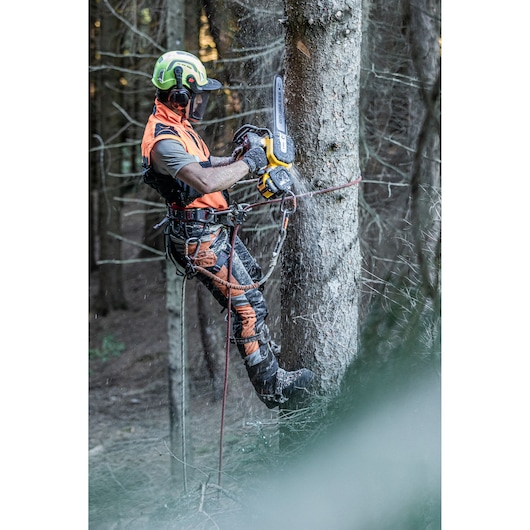 A person equipped with safety gear uses a DEWALT chainsaw to cut a tree while secured with climbing ropes in a forest.