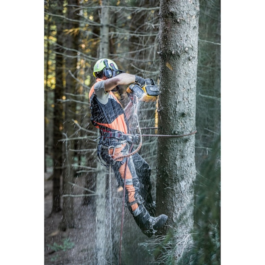 A person wearing safety gear uses a DEWALT chainsaw to cut a tree while suspended by ropes in a forest.