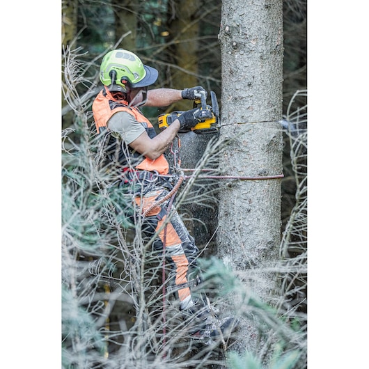 A person in safety gear, including a helmet and ear protection, is using a DEWALT chainsaw to cut a tree in a forest. The