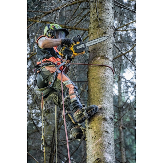 A person equipped with safety gear and climbing equipment uses a DEWALT cordless chainsaw to cut branches while climbing a