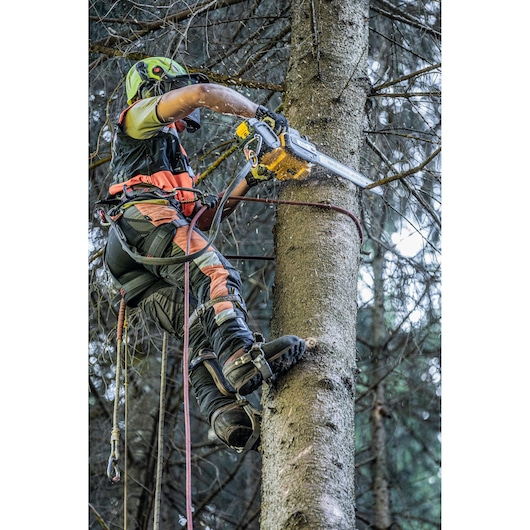 A person wearing safety gear and climbing equipment uses a DEWALT chainsaw to cut branches from a tree in a forest.