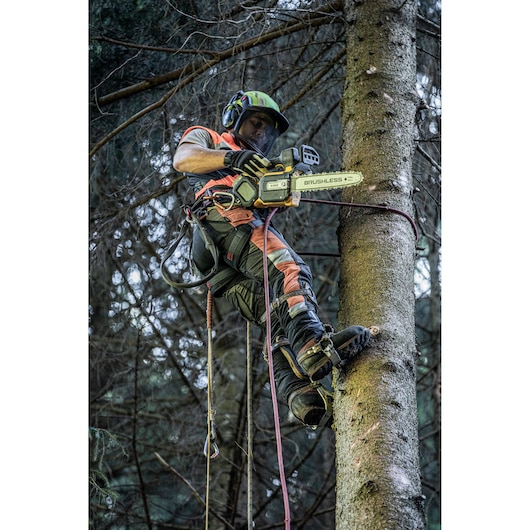 A person wearing safety gear and climbing equipment is using a DEWALT brushless chainsaw to cut branches from a tree. The