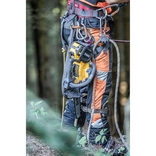 DEWALT chainsaw attached to a worker’s harness, worn over safety gear in a forest setting.