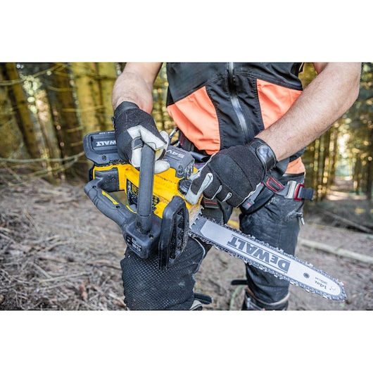 Person holding a DEWALT cordless chainsaw in a forest, wearing protective gloves and gear.