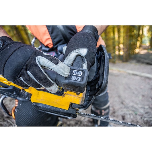 Gloved hands insert battery into yellow DEWALT chainsaw outdoors. Forest background visible.
