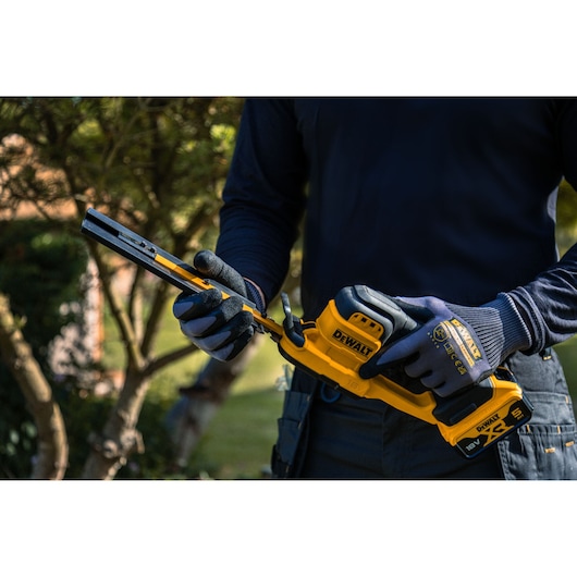 A person wearing gloves holds a DEWALT cordless hedge trimmer outdoors, with trees in the background.