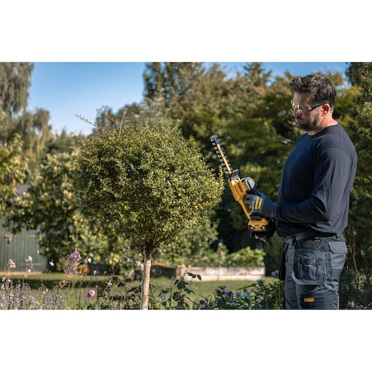 A person using a DEWALT cordless hedge trimmer (model DCMHT520N-XJ) to trim a rounded bush in a garden. The person’s face is blurred for privacy. The scene shows greenery and flowers under a clear sky.