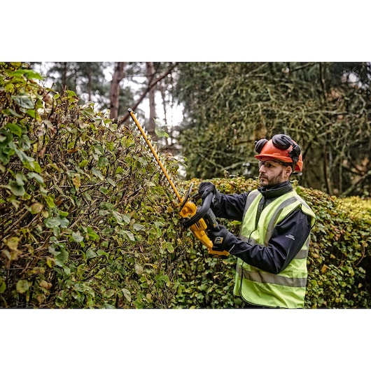 A person wearing safety gear and a high-visibility vest is using a DEWALT cordless hedge trimmer to trim a hedge outdoors.