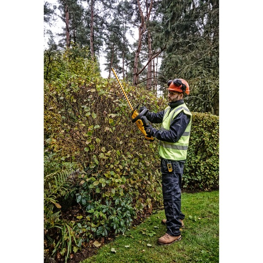 A person wearing safety gear and a high-visibility vest is using a DEWALT cordless hedge trimmer (model DCMHT563N) to trim bushes in a garden setting.