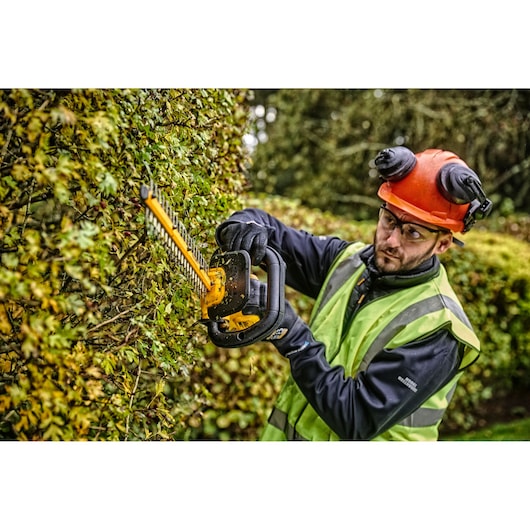 A person wearing safety gear, including an orange helmet and a reflective vest, uses a DEWALT DCMHT563N hedge trimmer to cut hedges outdoors. The hedge trimmer is yellow and black and designed for professional gardening and landscape maintenance.