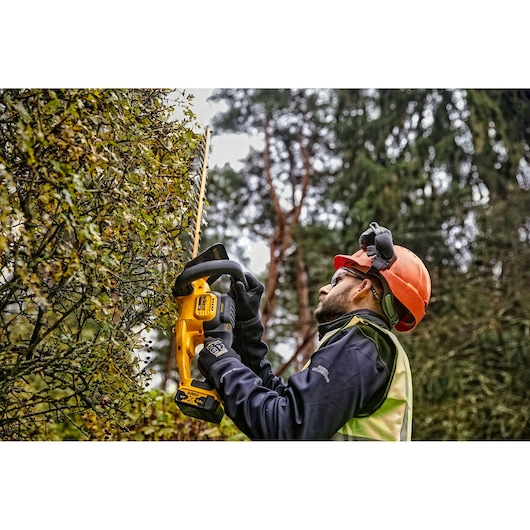 A person wearing protective gloves, helmet, and a high-visibility vest is using a yellow DEWALT cordless hedge trimmer to cut a hedge outdoors in a wooded area.