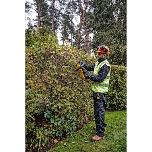 A person wearing safety gear and a high-visibility vest is using a DEWALT cordless hedge trimmer to trim a large bush in a garden.