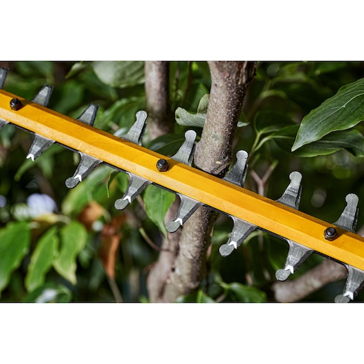 Close-up of a DEWALT hedge trimmer blade cutting through a tree branch with green leaves in the background.