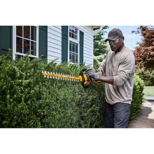 A person wearing gloves and a cap is using a DEWALT cordless hedge trimmer to trim bushes outside a house.